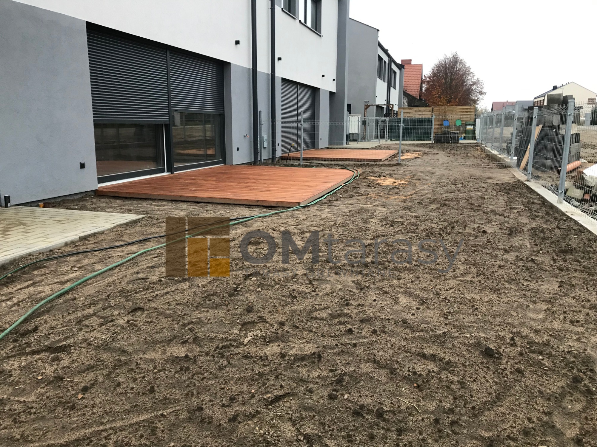 Dwa tarasy z desek kompozytowych przed nowoczesnym budynkiem w trakcie prac wykończeniowych ogrodu, widoczne ogrodzenie i przygotowany teren pod trawnik.