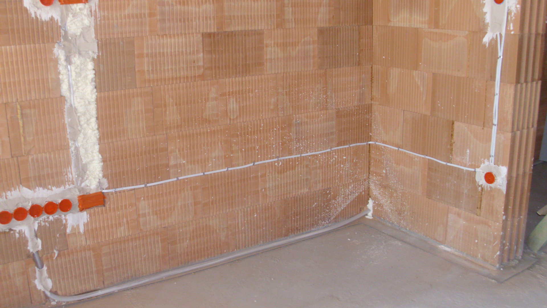 Ściana z pustaków ceramicznych z widocznymi przewodami elektrycznymi, puszkami instalacyjnymi w kolorze pomarańczowym i pianką montażową uszczelniającą bruzdy.