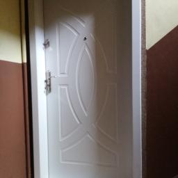 Km-Dre - Białe drzwi wejściowe z dekoracyjnym wzorem i srebrnymi okuciami, widoczne na tle ściany w dwóch odcieniach brązu i beżu.