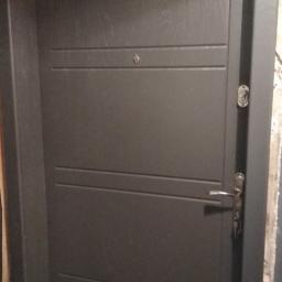 Km-Dre - Ciemnoszare drzwi z trzema poziomymi wgłębieniami, zamontowane w surowym otworze z widocznym betonem i resztkami zaprawy.