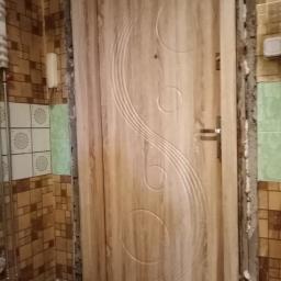 Km-Dre - Jasne drewniane drzwi wewnętrzne z dekoracyjnym frezem w kształcie okręgów i fal, widoczne w trakcie montażu, z resztkami zaprawy na framudze i fragmentem łazienki w tle.