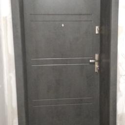 Km-Dre - Ciemnoszare drzwi antywłamaniowe z trzema poziomymi frezowaniami, wizjerem i zamkiem w ościeżnicy w trakcie montażu, widoczne ślady zaprawy na ścianie.