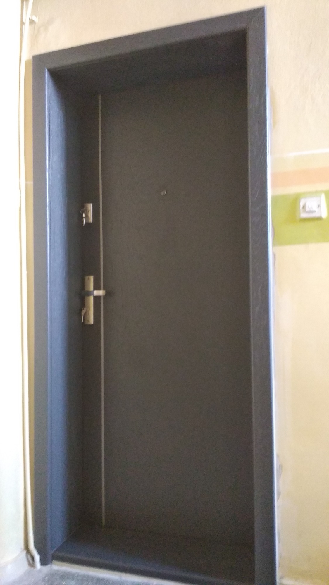 Szare drzwi wejściowe z widocznym zamkiem i kluczami, osadzone w ościeżnicy, na tle ściany z żółtą i zieloną farbą.