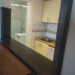 P.H.U. REMONT Andrzej Staniszewski - Fragment kuchni z widokiem na łazienkę z kabiną prysznicową, widoczne żółte szafki kuchenne, zlew i pralka, całość oddzielona blatem z ciemnego drewna.