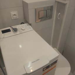 P.H.U. REMONT Andrzej Staniszewski - Pralka Indesit ustawiona obok toalety w małej łazience z szarymi płytkami, widoczny przycisk spłuczki i gniazdko elektryczne.