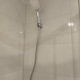 P.H.U. REMONT Andrzej Staniszewski - Nowoczesna, kwadratowa słuchawka prysznicowa z chromowanym wykończeniem na tle jasnych, beżowych płytek w kabinie prysznicowej.