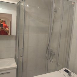 P.H.U. REMONT Andrzej Staniszewski - Wnętrze łazienki z kabiną prysznicową z zaokrąglonymi szybami i srebrnymi profilami, umywalką z szafką oraz pralką Indesit. Widoczne odbicie osoby robiącej zdjęcie w lustrze.