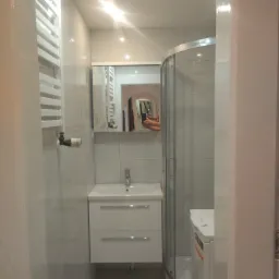 Niewielka, jasna łazienka z kabiną prysznicową, umywalką z szafką i grzejnikiem drabinkowym. W lustrze odbija się osoba robiąca zdjęcie telefonem.