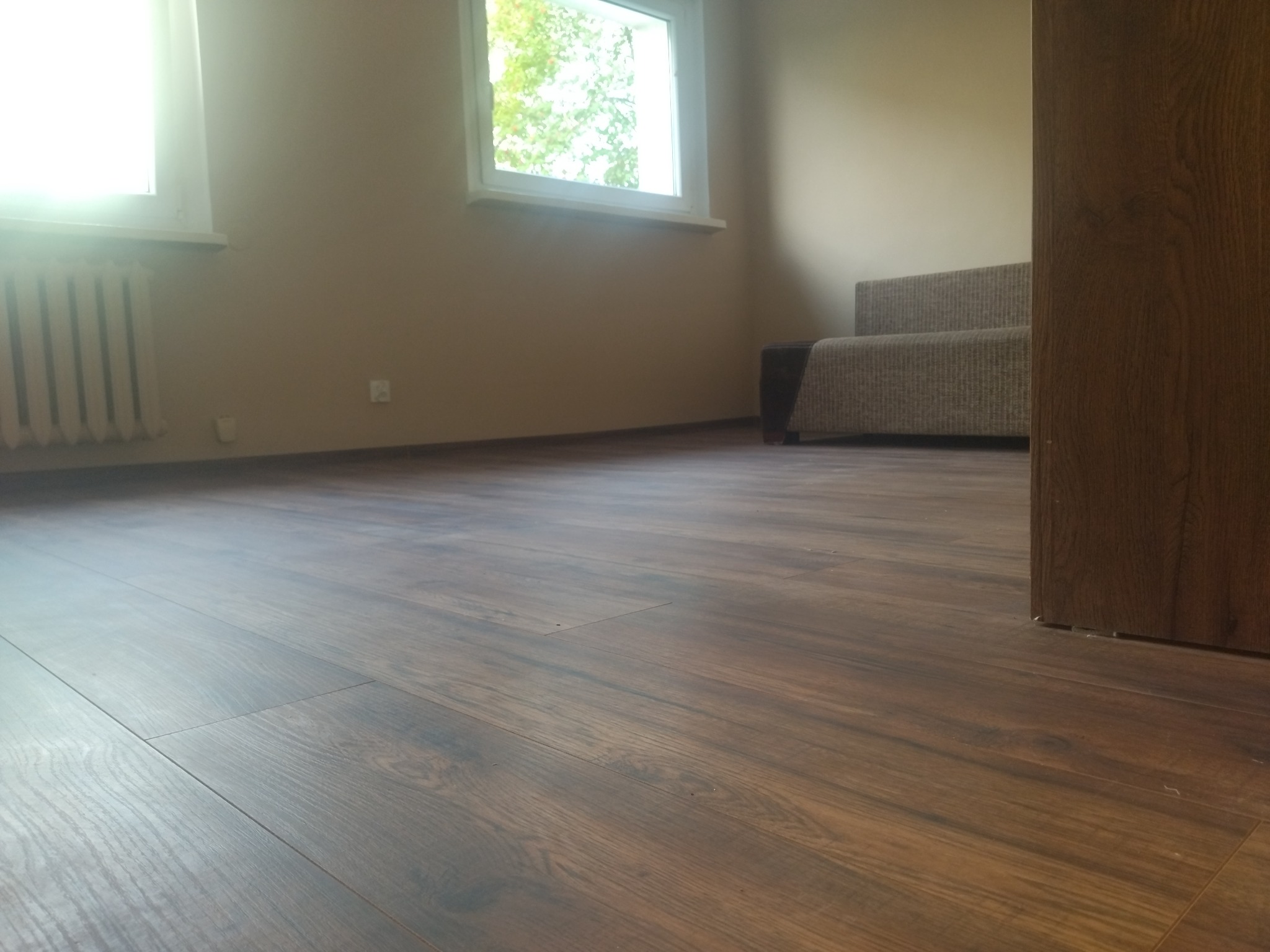Nowa, brązowa podłoga laminowana w jasnym pokoju z grzejnikiem i oknem, widziana z niskiego kąta, z fragmentem sofy i drzwi w tle.