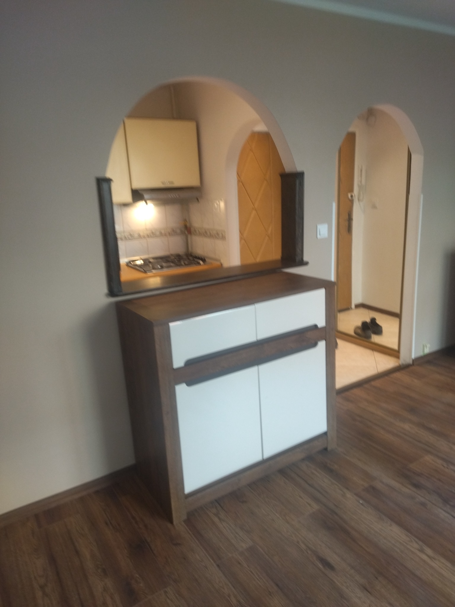 Aranżacja wnętrza z szafką z białymi frontami i drewnianym blatem, widok na kuchnię przez łukowate przejście, w tle wejście do innego pomieszczenia.