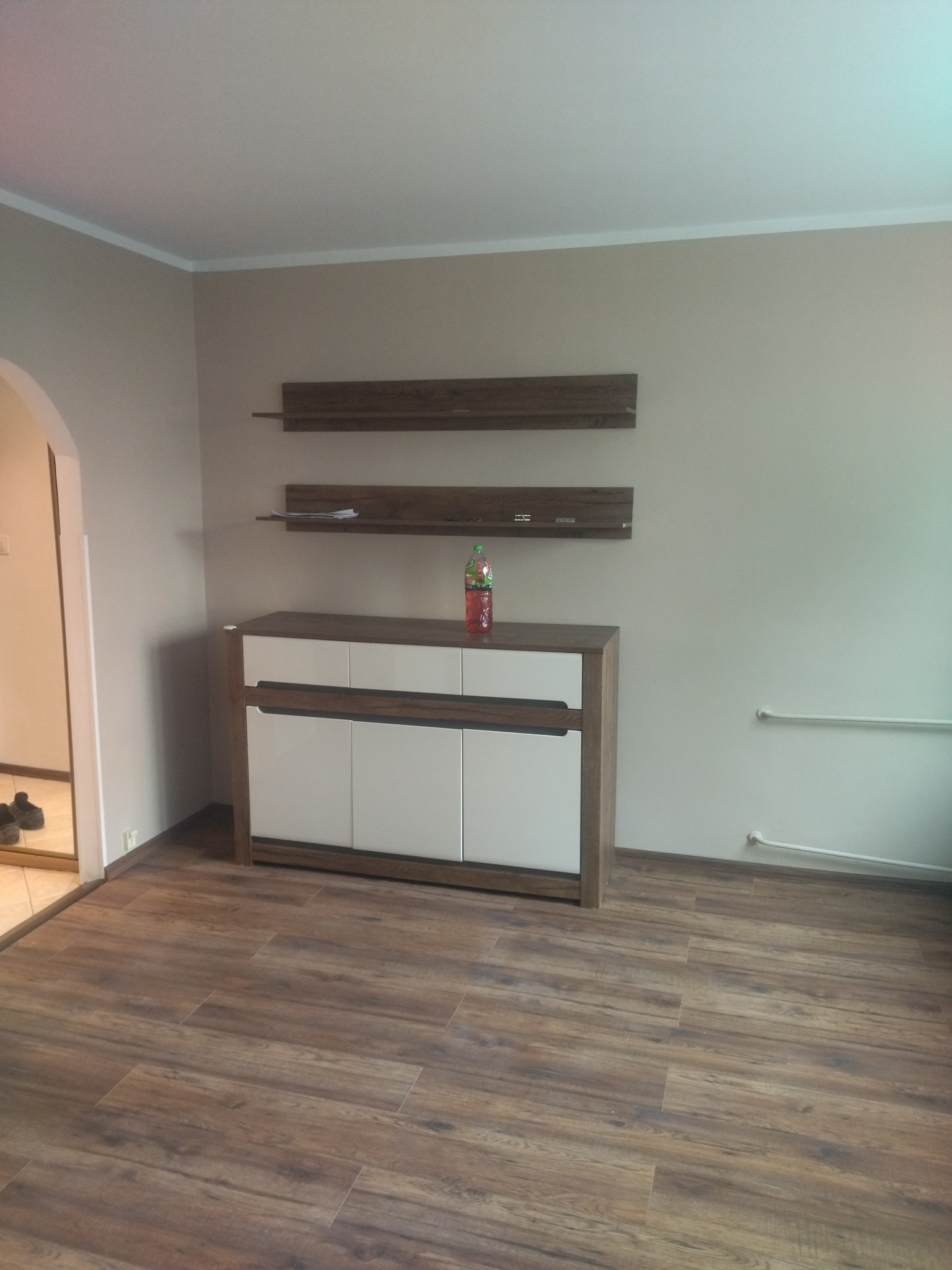 Nowoczesny salon z brązową podłogą imitującą drewno, białą komodą z brązowymi akcentami i dwiema półkami wiszącymi na ścianie w kolorze beżowym, na komodzie stoi butelka napoju.