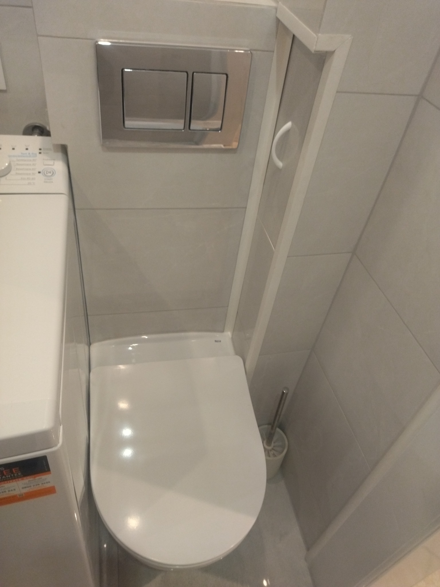 Kompaktowa toaleta wisząca z przyciskiem spłukującym w chromowanej ramce, obok pralka w minimalistycznej łazience wykończonej szarymi płytkami.
