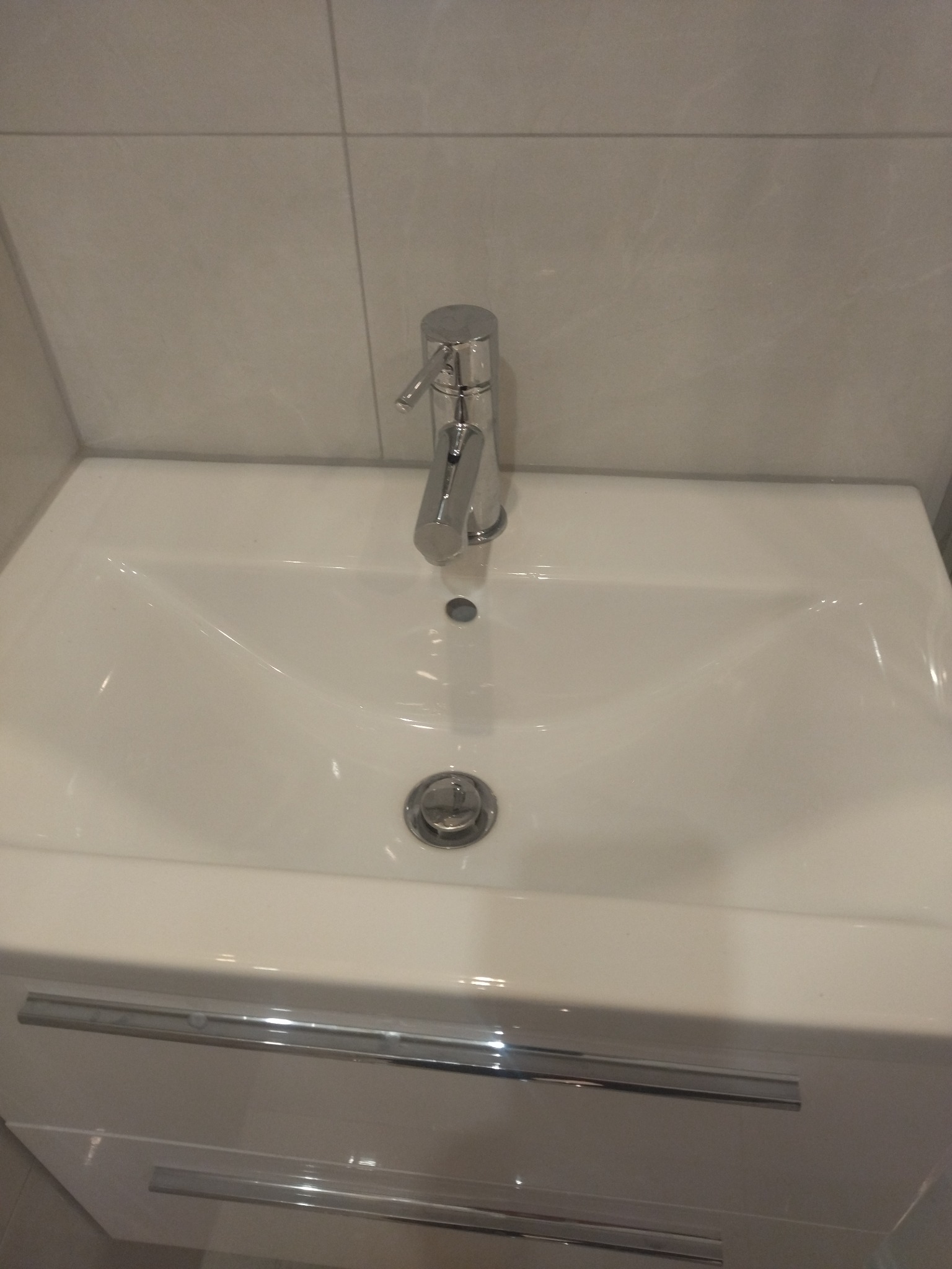 Biała umywalka z szafką i chromowaną baterią na tle jasnych płytek w łazience.