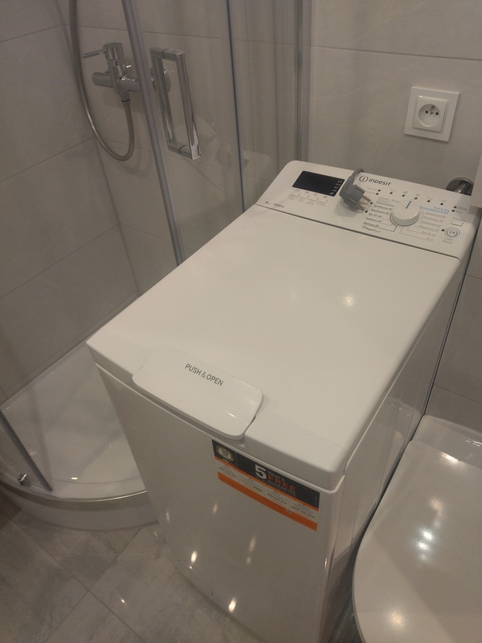 Nowa pralka marki Indesit z górnym załadunkiem w łazience, widoczna obok kabiny prysznicowej i toalety, z naklejką o 5-letniej gwarancji.