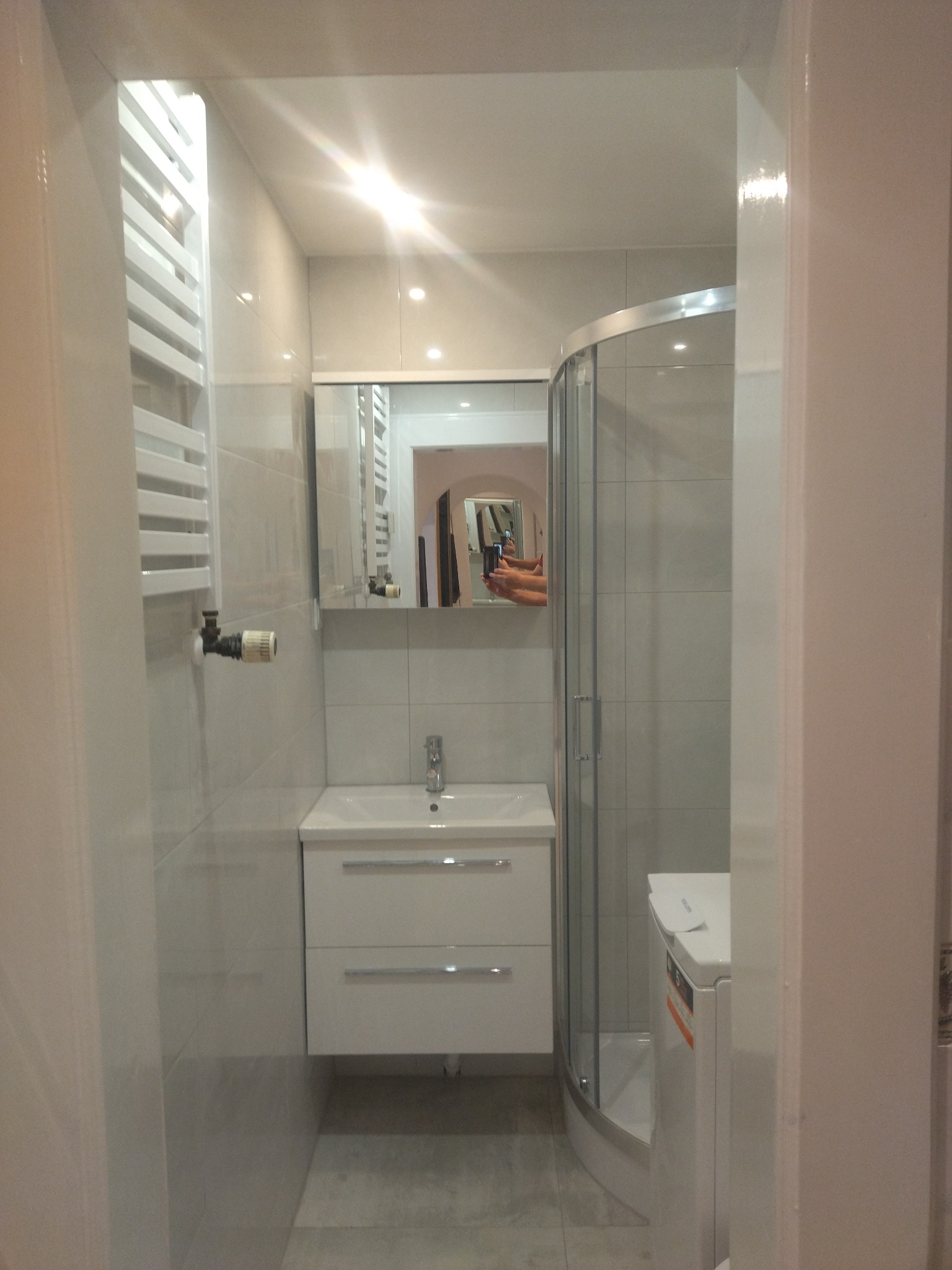 Niewielka, jasna łazienka z kabiną prysznicową, umywalką z szafką i grzejnikiem drabinkowym. W lustrze odbija się osoba robiąca zdjęcie telefonem.