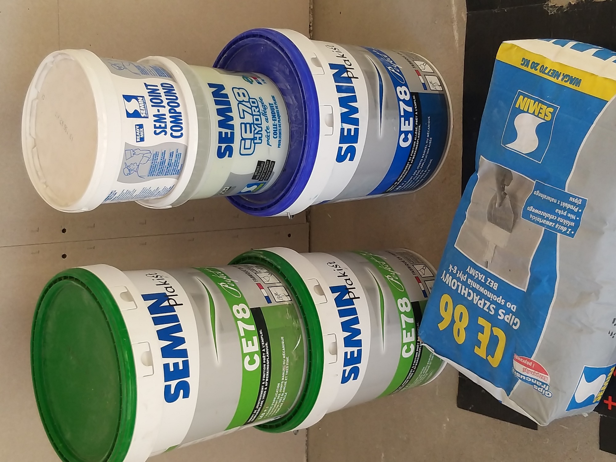 Ułożone obok siebie opakowania z masami szpachlowymi i gipsowymi marki Semin: wiadro z masą do spoinowania płyt g-k, wiadro z gładzią szpachlową i worek z gipsem szpachlowym.