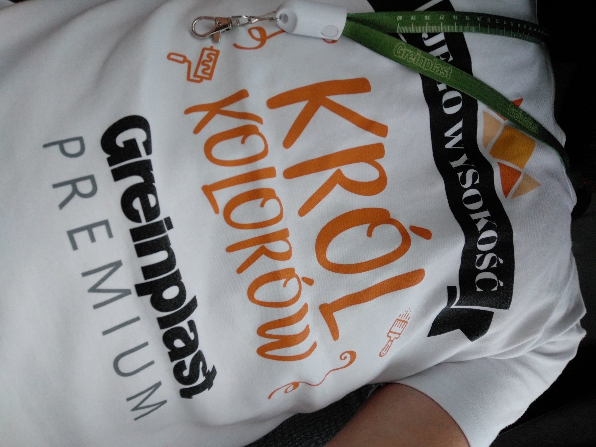 Biała koszulka z nadrukiem 'Król Kolorów' z logo firmy Greinplast, widoczny fragment smyczy z miarką zwiniętą w kółko.