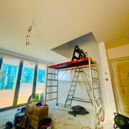 Usługi Remontowo Wykończeniowe PIOTR LEONOWICZ - Wnętrze w trakcie malowania: aluminiowy ruszt LEMAR, zabezpieczone okna folią, puszki z farbą Dulux. Widoczny fragment pomalowanego sufitu.