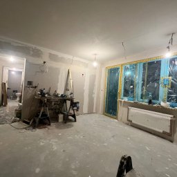 Usługi Remontowo Wykończeniowe PIOTR LEONOWICZ - Wnętrze w trakcie prac wykończeniowych: ściany przygotowane pod gładzie, okna zabezpieczone folią, widoczne narzędzia i drabina. Podłoga pokryta warstwą wyrównującą.