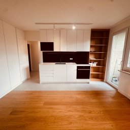 Usługi Remontowo Wykończeniowe PIOTR LEONOWICZ - Nowoczesna kuchnia z białymi szafkami, czarnym panelem nad blatem i drewnianymi półkami. Parkiet w odcieniach brązu, duże okno z widokiem na zieleń. Minimalistyczny design.