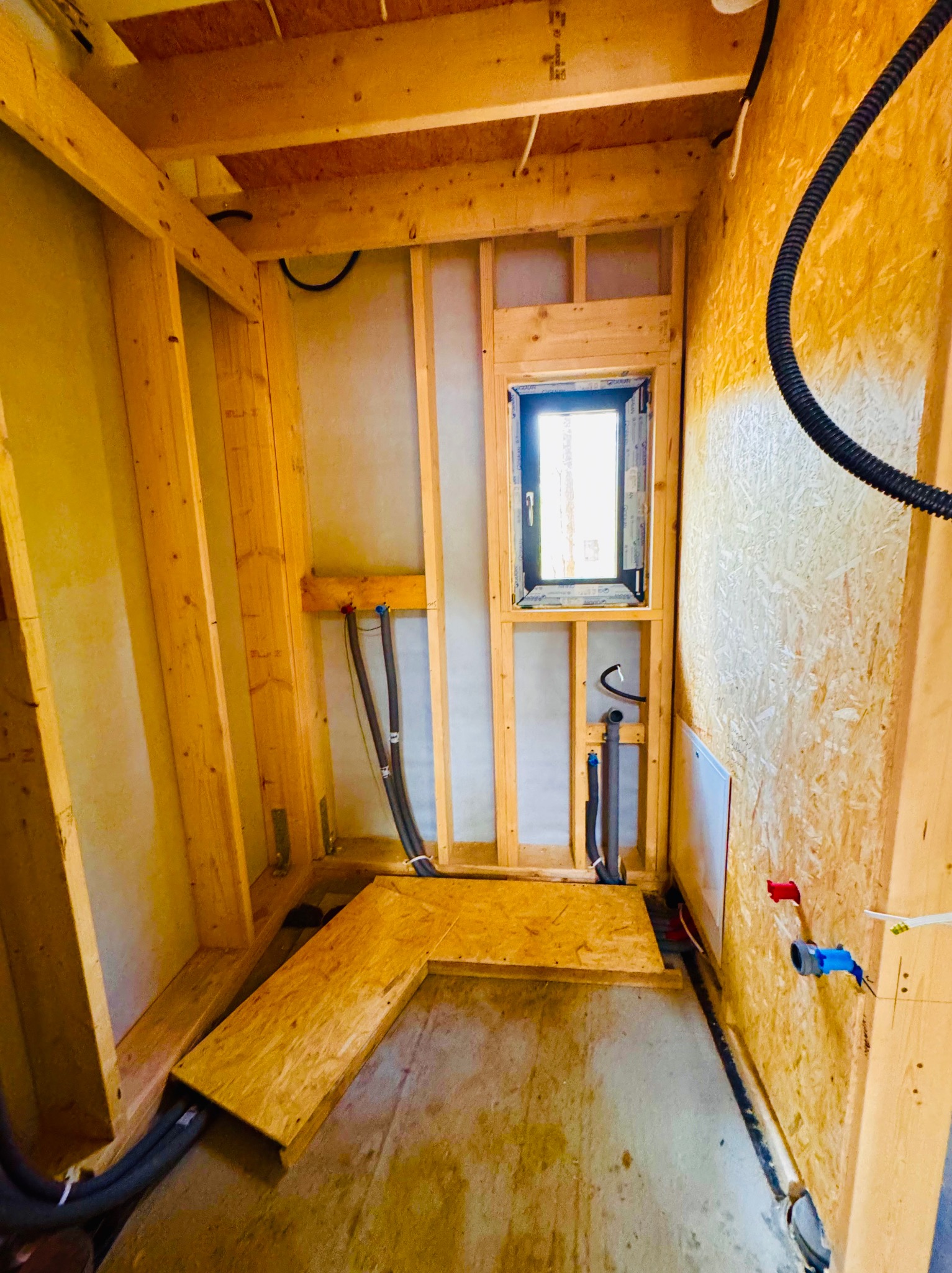 Wnętrze w trakcie budowy: drewniana konstrukcja szkieletowa, okno z czarną ramą, instalacje wodne i elektryczne, płyta OSB na ścianie. Surowy stan wykończenia.