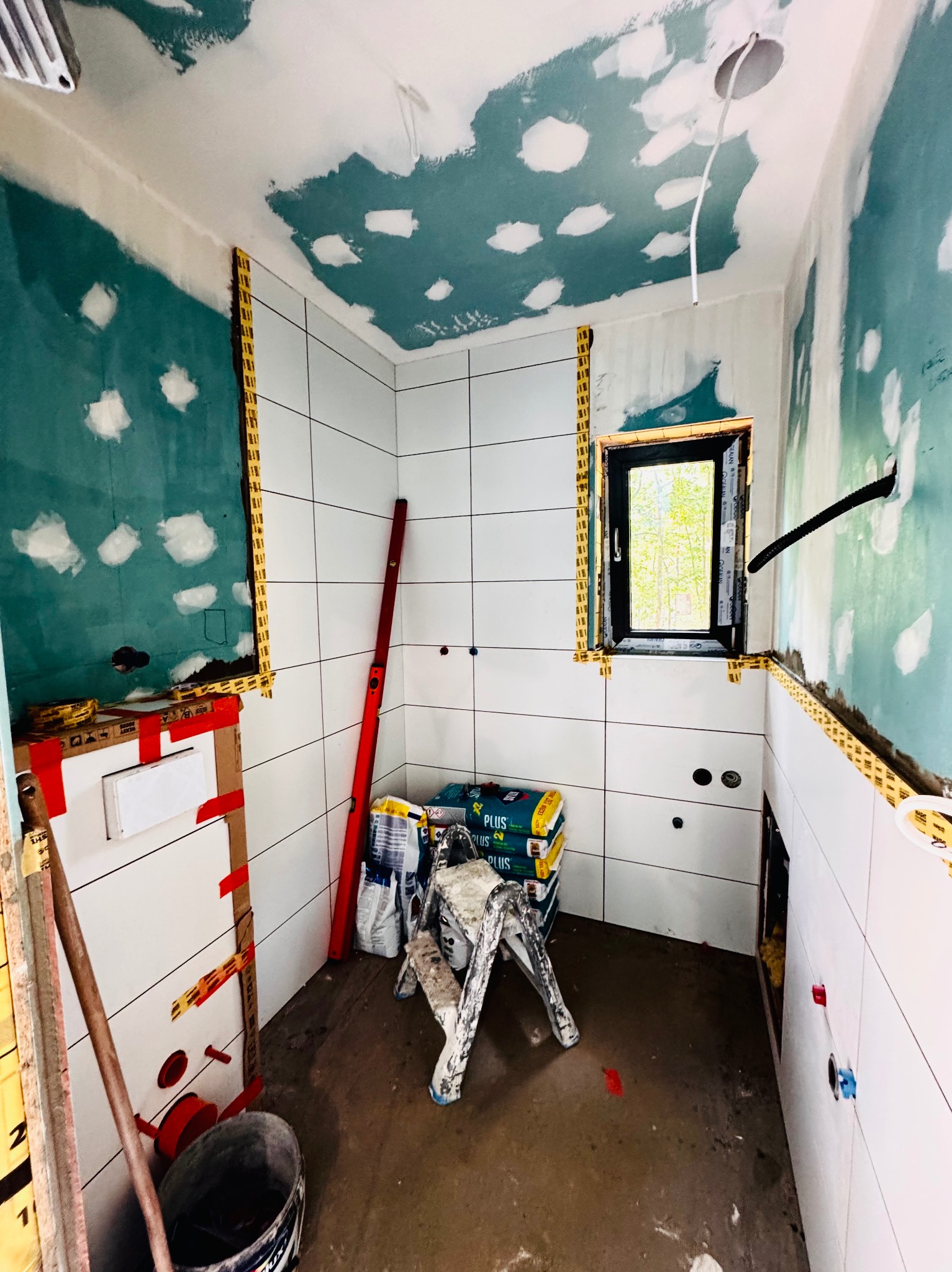 Wnętrze łazienki w trakcie remontu: białe płytki na ścianach, przygotowane ściany pod malowanie, okno z widokiem na zieleń, narzędzia i materiały budowlane.