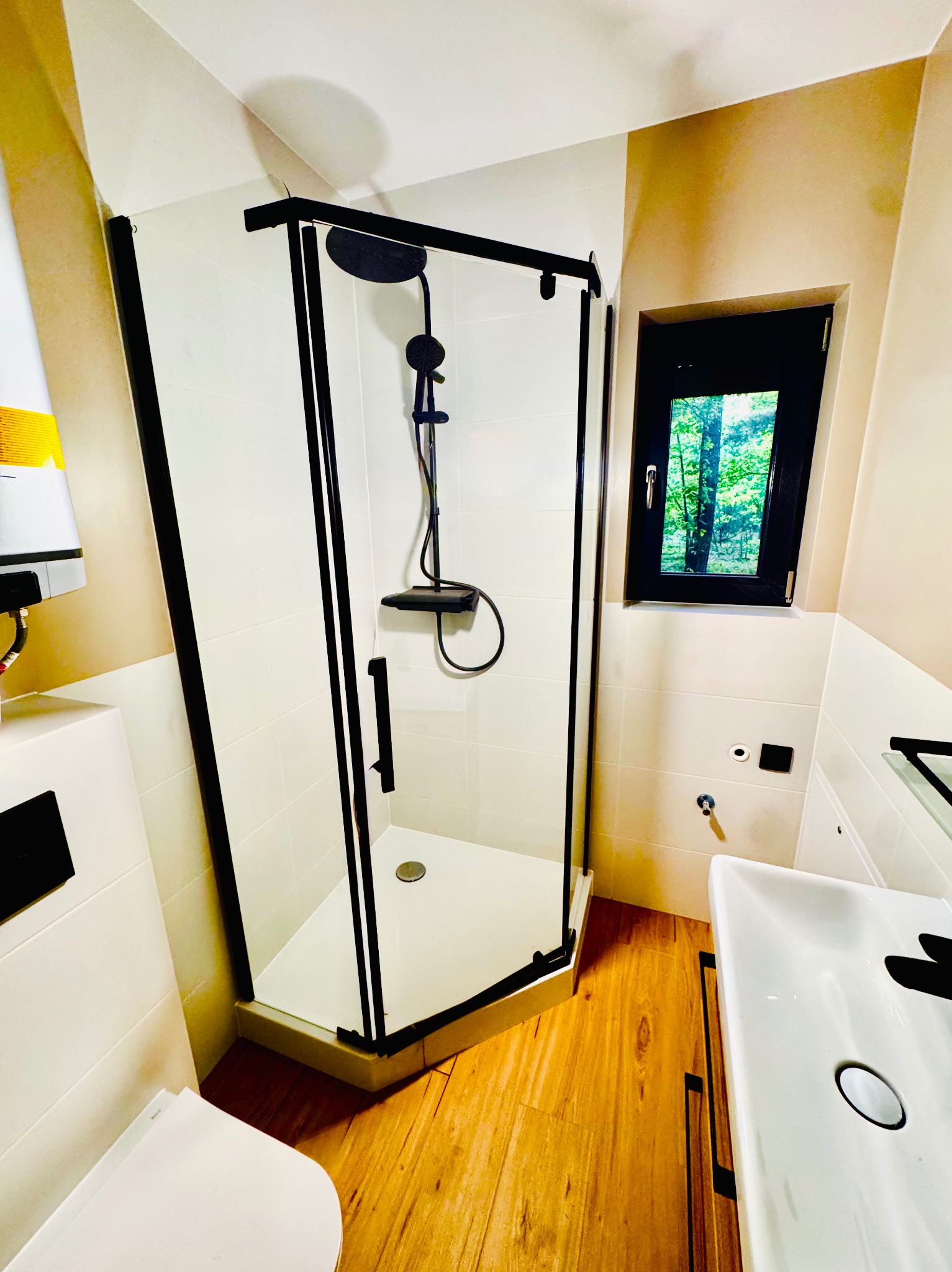 Nowoczesna kabina prysznicowa z czarnymi profilami, białe płytki, drewniana podłoga i okno z widokiem na zieleń. Minimalistyczny design łazienki po remoncie.