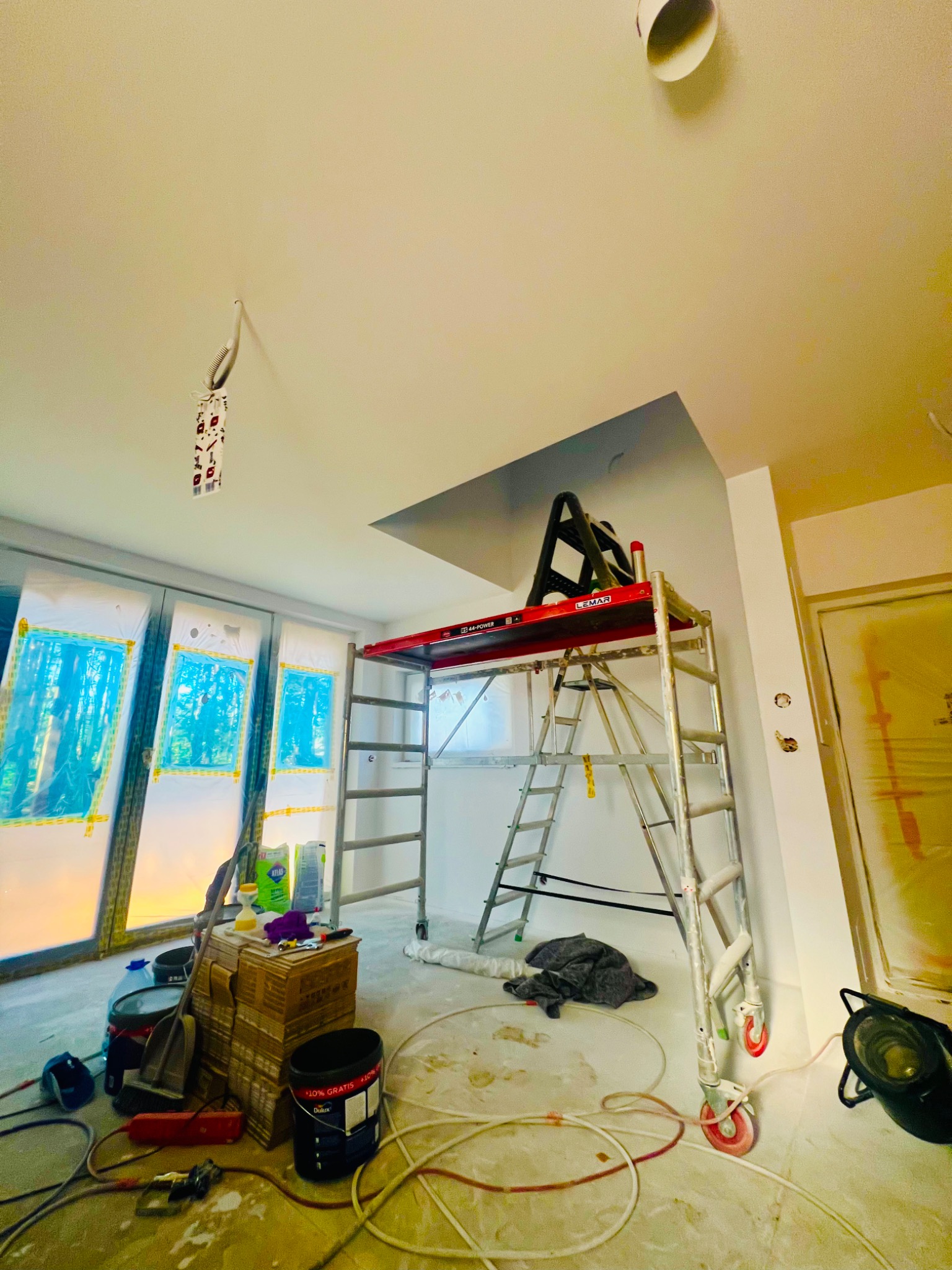 Wnętrze w trakcie malowania: aluminiowy ruszt LEMAR, zabezpieczone okna folią, puszki z farbą Dulux. Widoczny fragment pomalowanego sufitu.