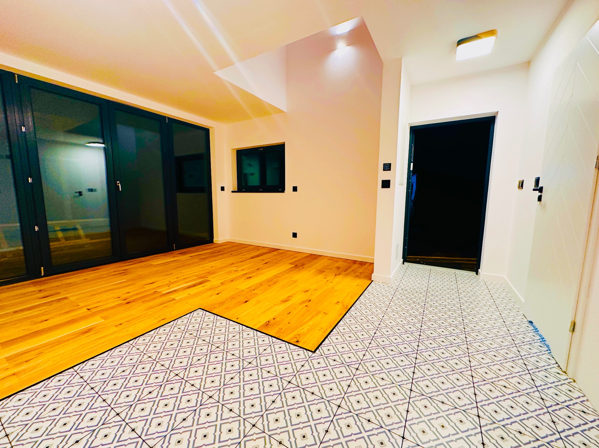 Jasne wnętrze z parkietem i wzorzystą podłogą w korytarzu. Widoczne duże okna balkonowe i drzwi. Kontrast materiałów i kolorów. Nowoczesne wykończenie.