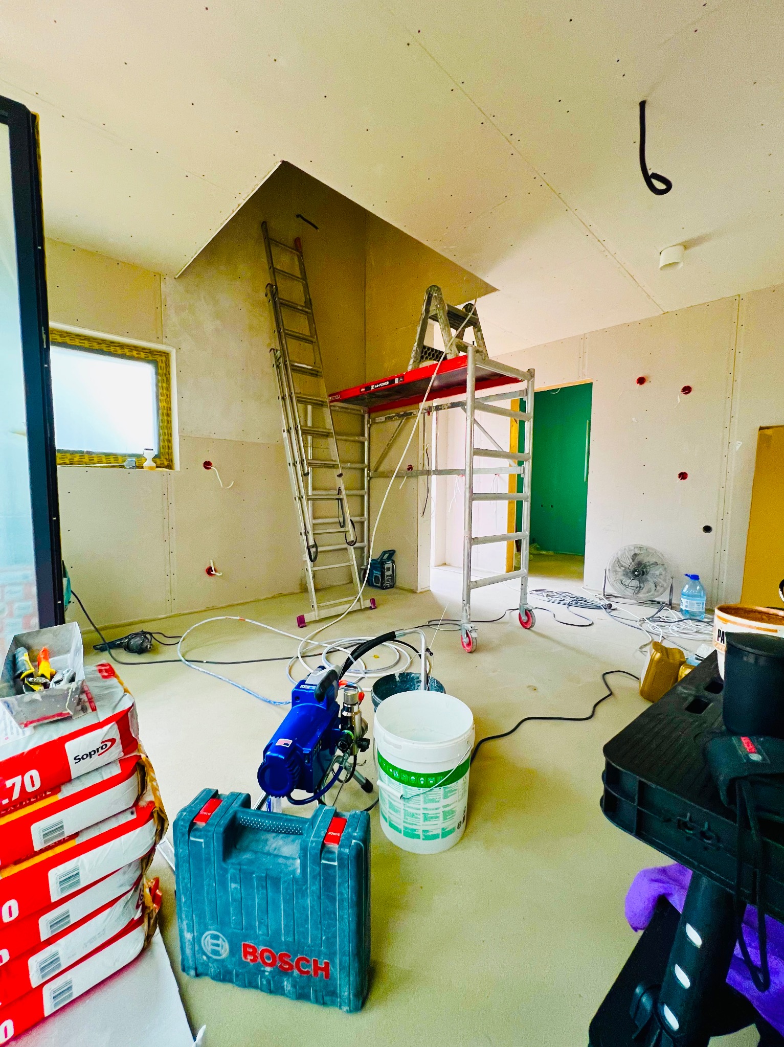 Wnętrze w trakcie prac wykończeniowych: ściany z płyt gipsowo-kartonowych, drabina, rusztowanie, narzędzia malarskie, worki z zaprawą. Widoczne otwory na instalacje.
