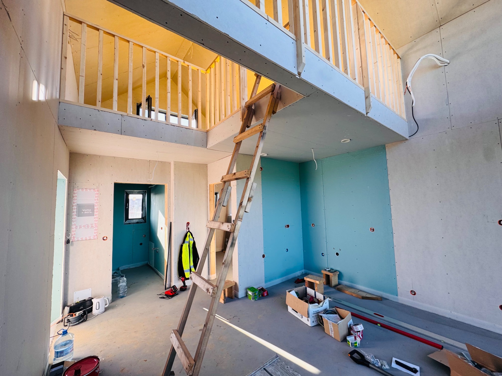 Wnętrze w trakcie montażu płyt gipsowo-kartonowych z drewnianą antresolą i schodami. Widoczne narzędzia i materiały budowlane. Ściany częściowo pomalowane na niebiesko.