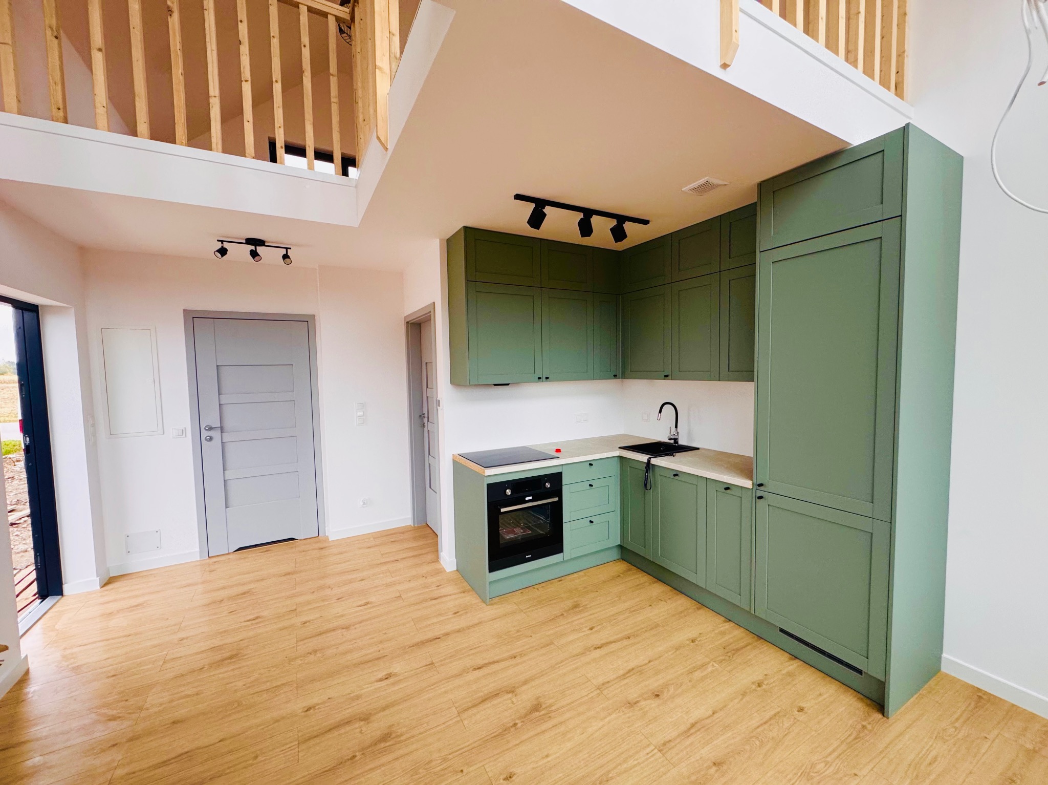 Nowoczesna kuchnia w odcieniach zieleni z drewnianą podłogą i antresolą. Minimalistyczne oświetlenie punktowe i szara stolarka drzwiowa.