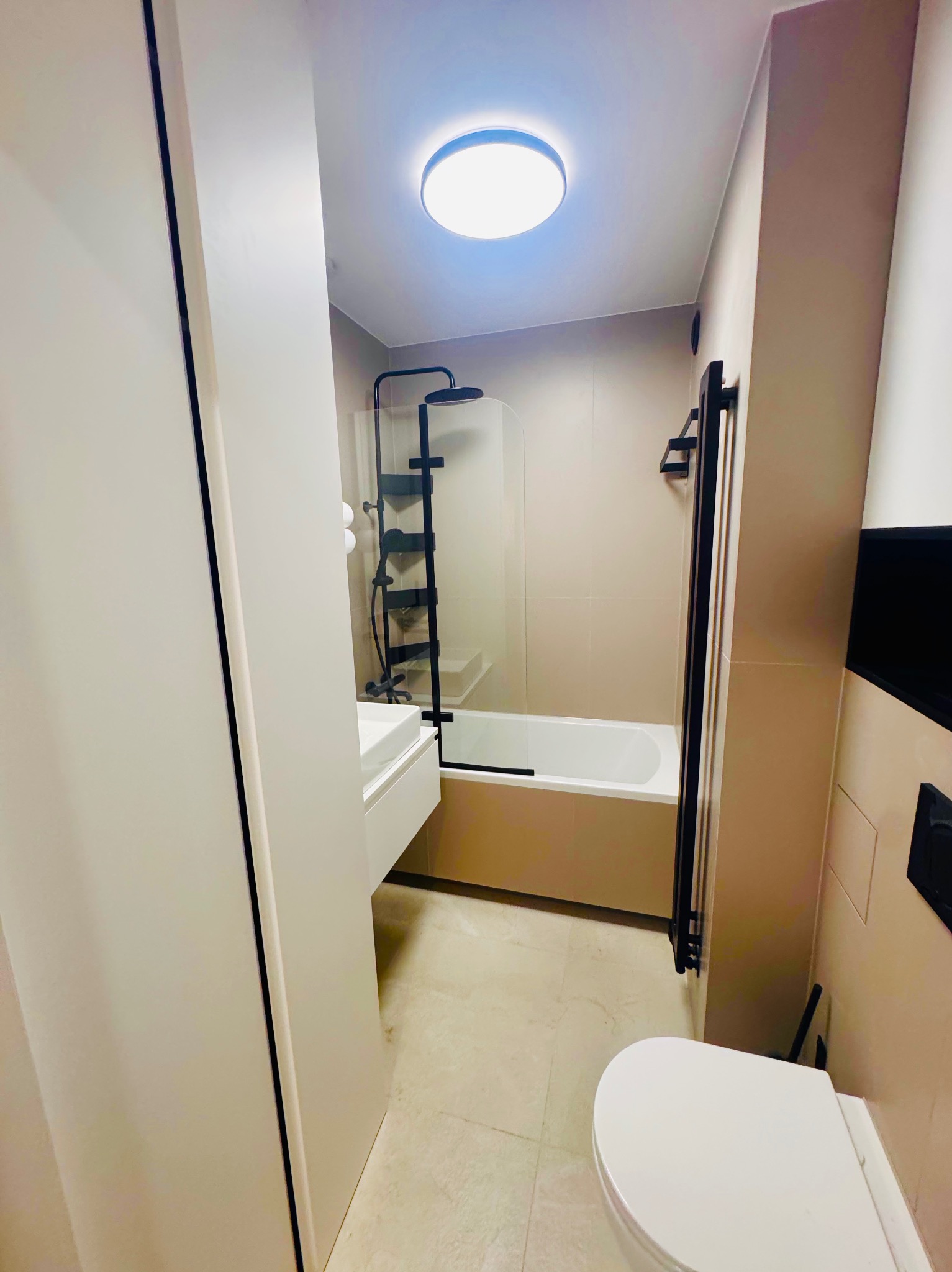 Nowoczesna łazienka po remoncie: beżowe płytki, biała armatura, czarne akcenty (grzejnik, rama prysznica). Minimalistyczny design, jasne oświetlenie.