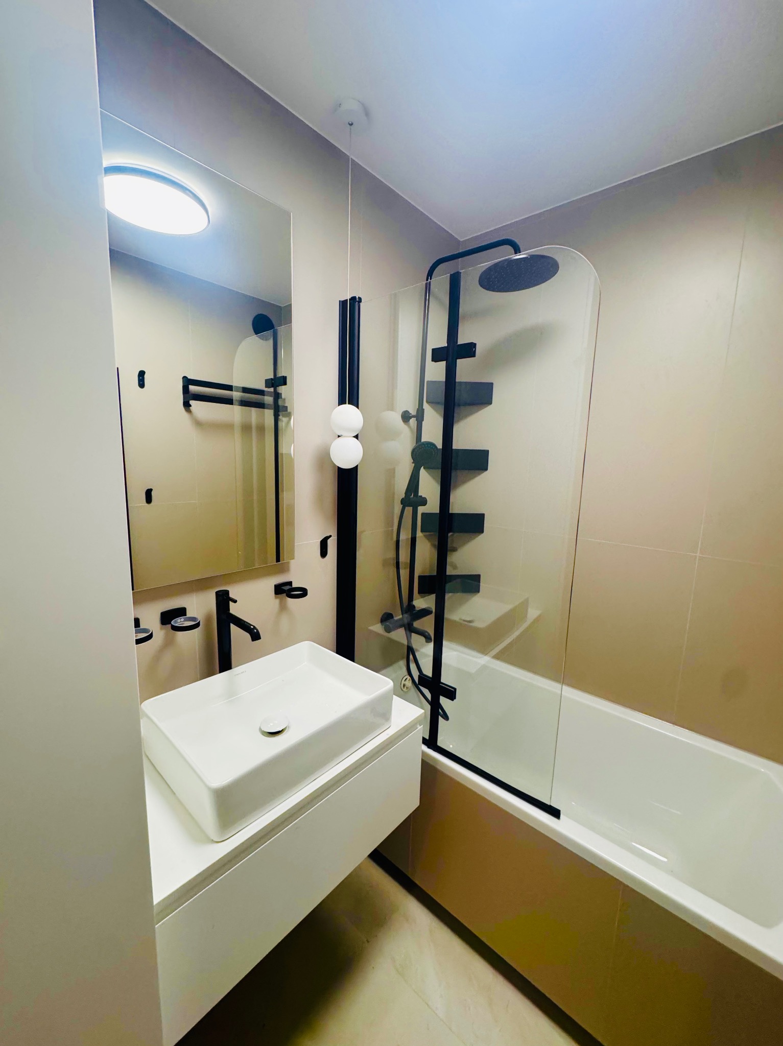 Nowoczesna łazienka z beżowymi płytkami, białą umywalką nablatową i czarną armaturą. Kabina prysznicowa z czarnymi elementami. Minimalistyczny design.