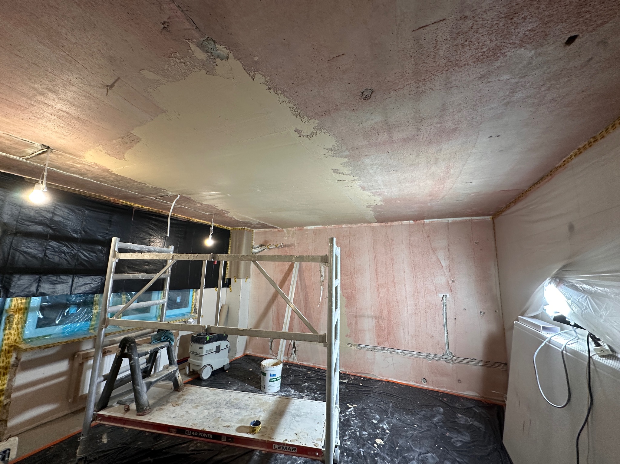 Pokój w trakcie prac wykończeniowych: częściowo wygładzony sufit, rusztowanie, zabezpieczone okno folią. Widoczne ślady po instalacjach elektrycznych na ścianach.