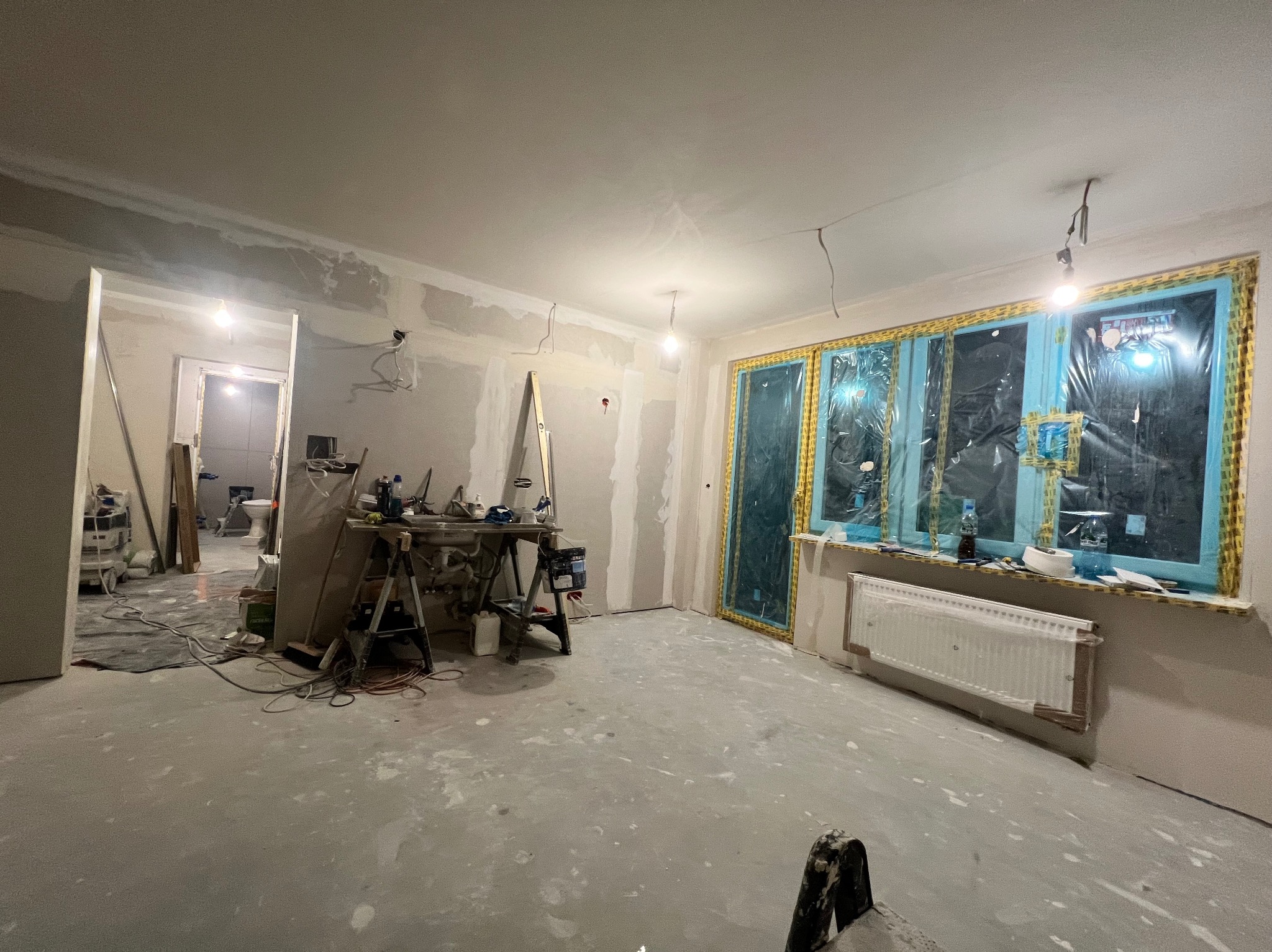 Wnętrze w trakcie prac wykończeniowych: ściany przygotowane pod gładzie, okna zabezpieczone folią, widoczne narzędzia i drabina. Podłoga pokryta warstwą wyrównującą.