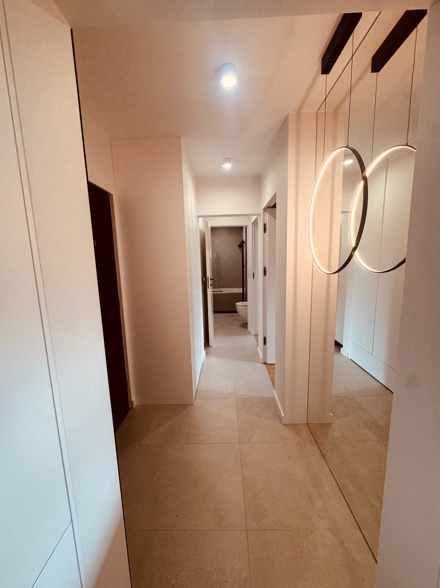 Nowoczesny korytarz z beżową podłogą, białymi ścianami i szafami w zabudowie. Okrągłe, wiszące lampy z ledowym oświetleniem. W tle widoczna łazienka.