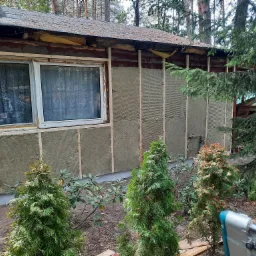 Ocieplenie ściany budynku z widoczną wełną mineralną i drewnianą konstrukcją, z przodu rosną krzewy iglaste