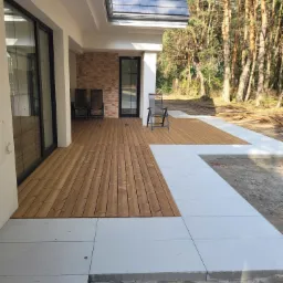 Nowoczesny taras drewniany z jasnymi płytami betonowymi, otoczony drzewami iglastymi, z widocznymi krzesłami ogrodowymi i fragmentem elewacji domu wykończonej cegłą.