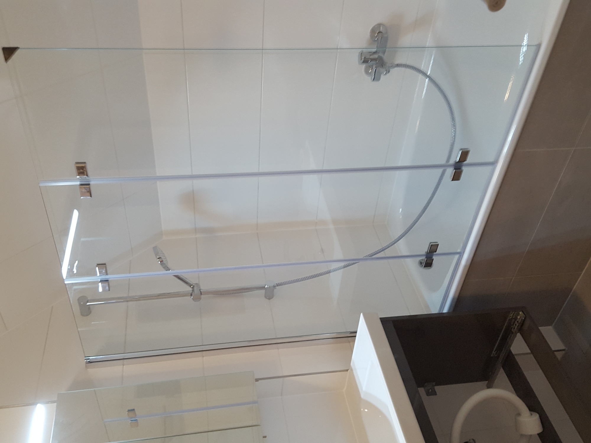Nowoczesna szklana ścianka prysznicowa zamontowana na wannie, z chromowanymi uchwytami i baterią, białe płytki na ścianie, szary pas płytek z boku, widoczna umywalka z szafką.
