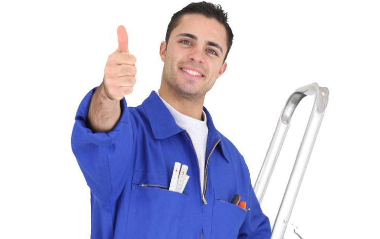 Uśmiechnięty mężczyzna w niebieskim kombinezonie roboczym, z miarką i narzędziami w kieszeniach, pokazuje kciuk w górę, stojąc obok aluminiowej drabiny na białym tle.