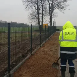 Pracownik w odblaskowej kurtce firmy 'Mi-Le-An Roboty Drogowe i Wodnokanalizacyjne' stoi tyłem obok nowo postawionego ogrodzenia z siatki panelowej na betonowej podmurówce, z łopatą w ręku, na tle...