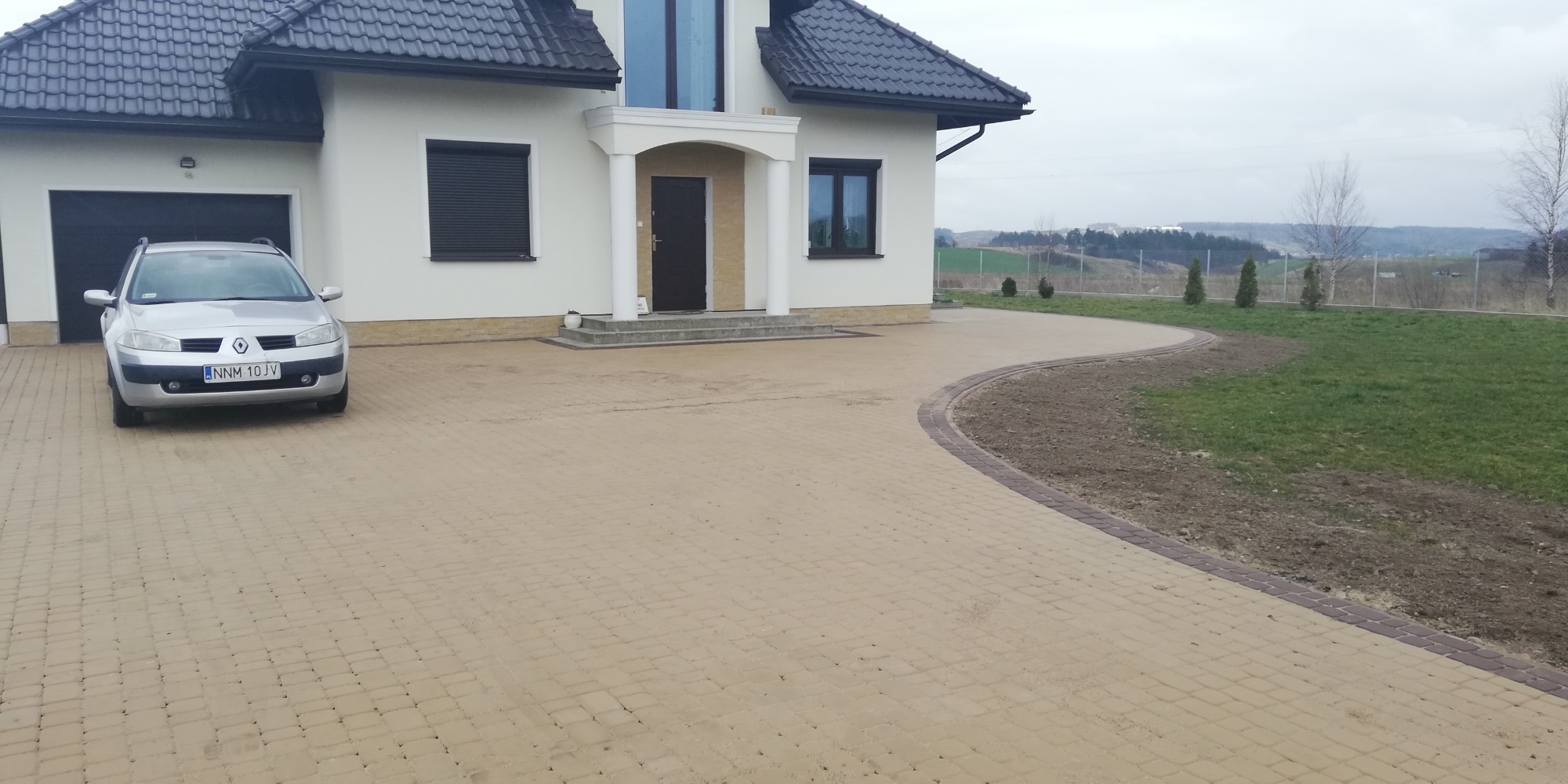 Ułożona kostka brukowa w Nowym Mieście Lubawskim tworzy podjazd do domu z widocznym samochodem i fragmentem ogrodu z młodymi drzewkami.