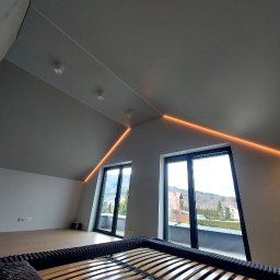 MAKTA Sp. z o.o. - Pokój na poddaszu z oknami balkonowymi i oświetleniem LED. Widoczne skosy, sufit w odcieniach szarości i fragment ramy łóżka. Nowoczesne wykończenie wnętrza.