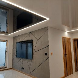 MAKTA Sp. z o.o. - Nowoczesne wnętrze po remoncie: błyszczący sufit z oświetleniem LED, ściana z geometrycznym wzorem, dwa telewizory i drewniane drzwi. Okna zabezpieczone folią.