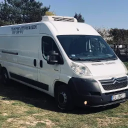 Biały samoch&oacute;d dostawczy z napisem 'TRANSPORT-PRZEPROWADZKI' i numerem telefonu, zaparkowany na trawie w słoneczny dzień.