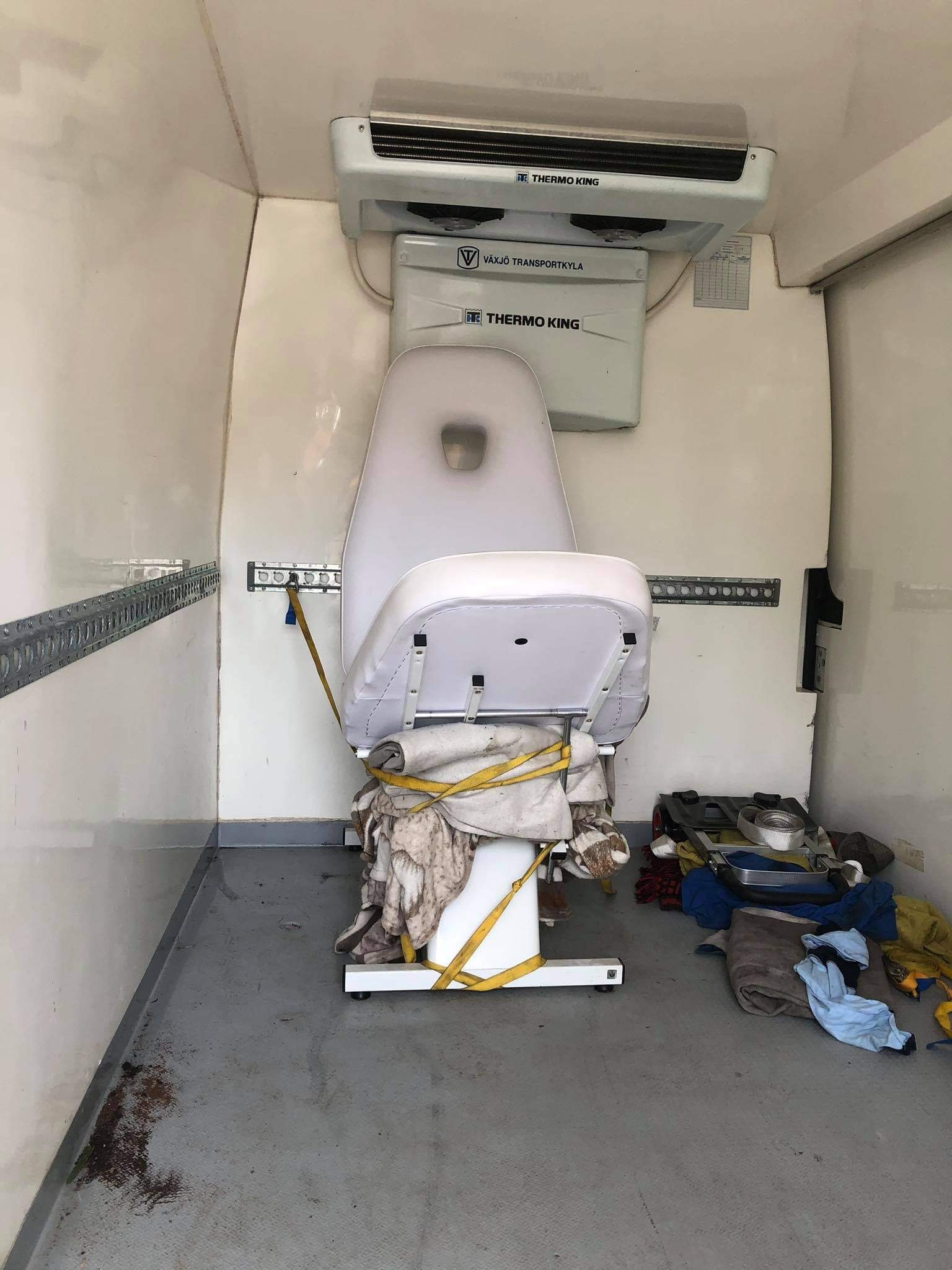 Wnętrze chłodni transportowej z zamocowanym, zabezpieczonym pasami fotelem medycznym i rozrzuconymi materiałami opatrunkowymi na podłodze.