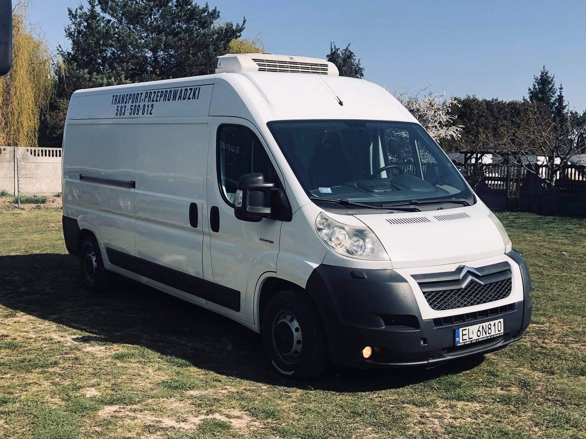 Biały samochód dostawczy z napisem 'TRANSPORT-PRZEPROWADZKI' i numerem telefonu, zaparkowany na trawie w słoneczny dzień.