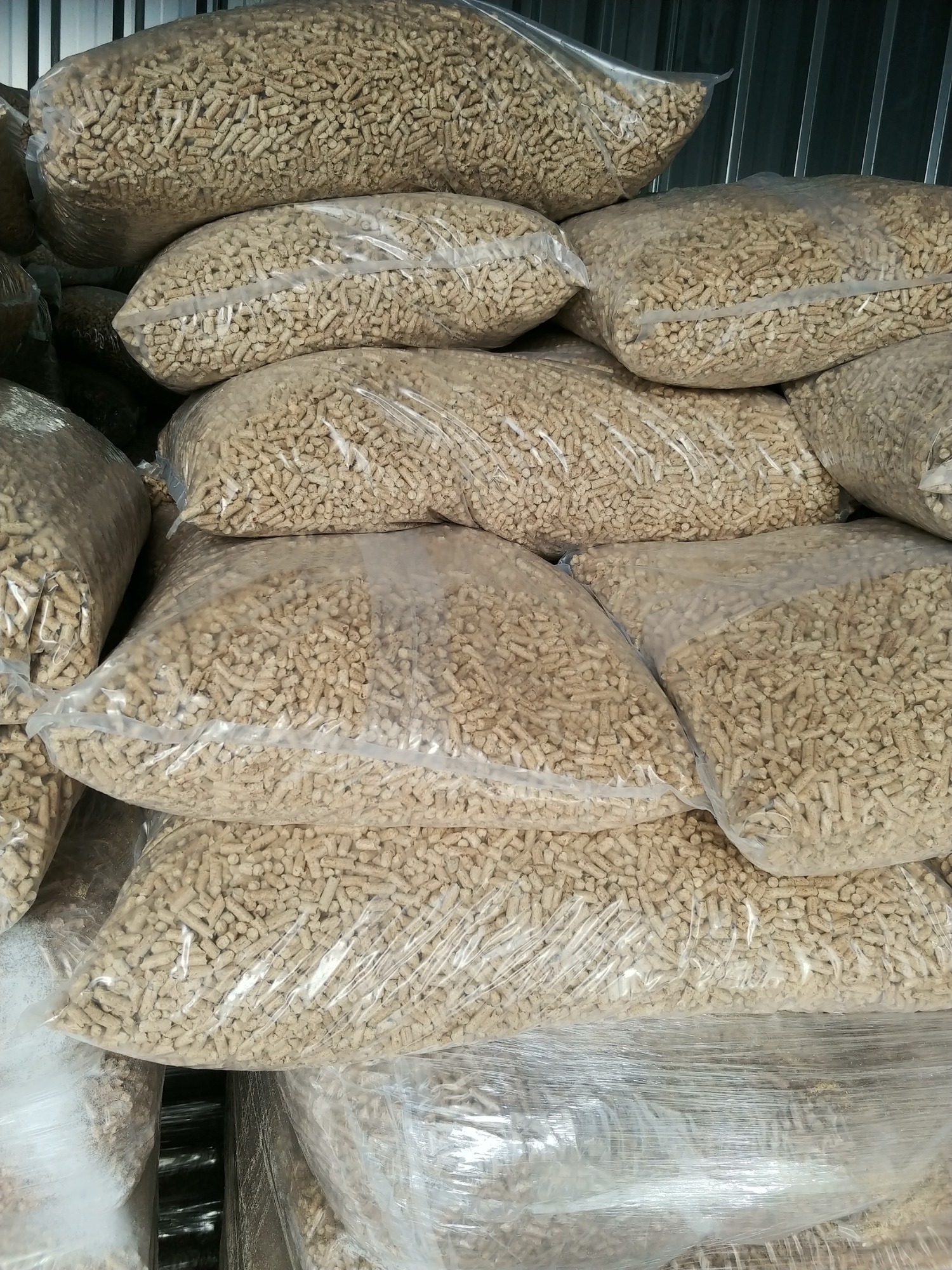 Stos worków wypełnionych jasnymi granulkami opału, zabezpieczonych folią, ułożonych w magazynie z blachy falistej.
