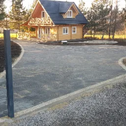Nowo ułożony, zakrzywiony podjazd z ciemnoszarej kostki brukowej z jasnymi obrzeżami prowadzący do drewnianego domu z balkonem, świeżo przygotowany teren pod trawnik po bokach, widoczne słupki...